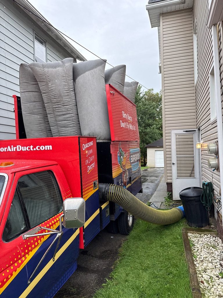 Superior duct-cleaning truck with gray filter bags up, parked in a narrow side yard with the yellow vacuum hose running to the house
