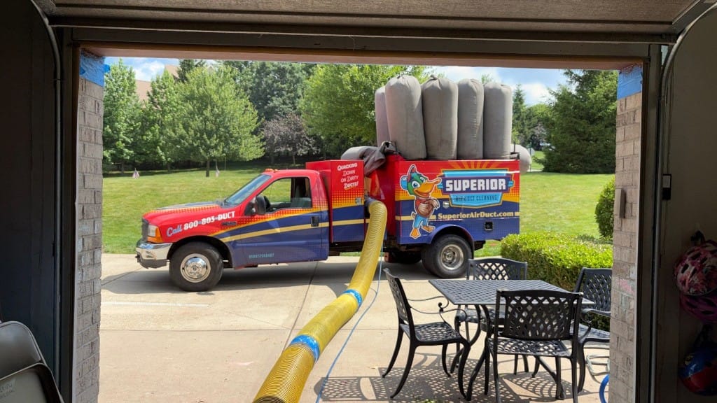 View through a customer's open garage: a Superior Air Duct Cleaning truck at the curb with the yellow negative-pressure vacuum hose running into the house