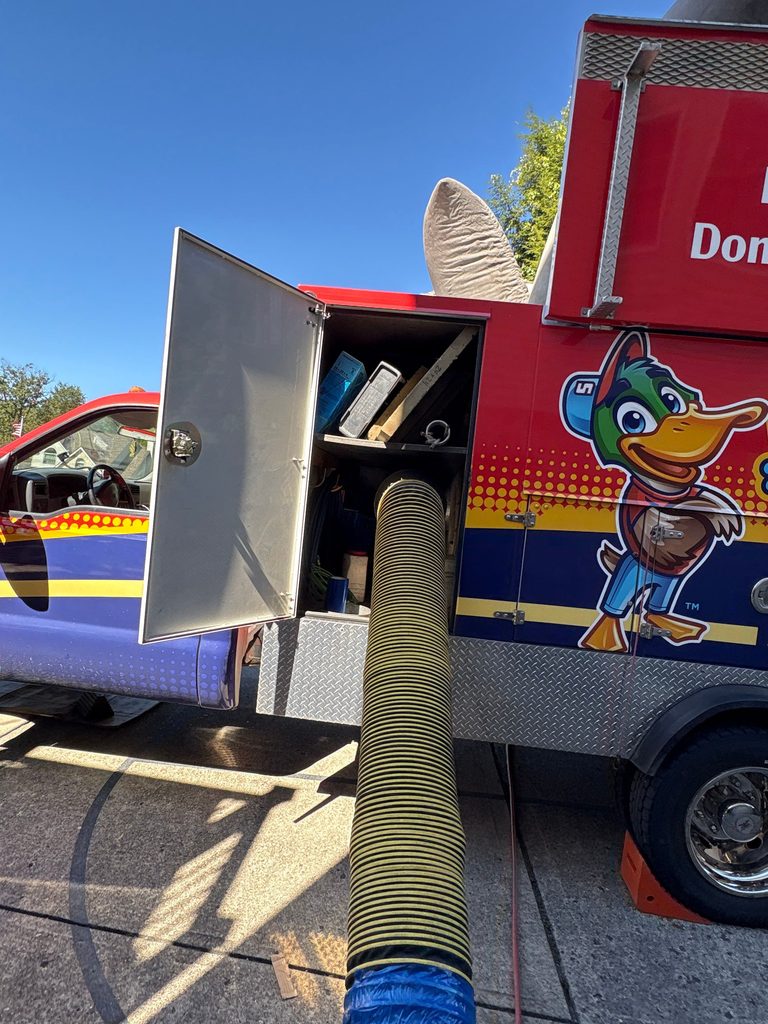 Yellow vacuum hose connected to the open equipment compartment of a Superior truck, next to the duck mascot wrap