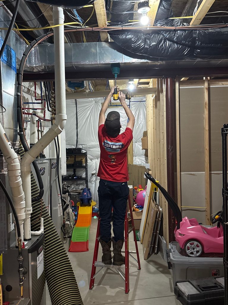 Superior technician on a ladder working at the basement ductwork, with the vacuum hose running across the floor