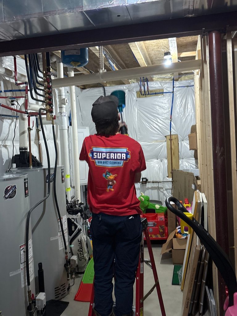 Superior technician in a red company shirt working at the basement ductwork beside the furnace