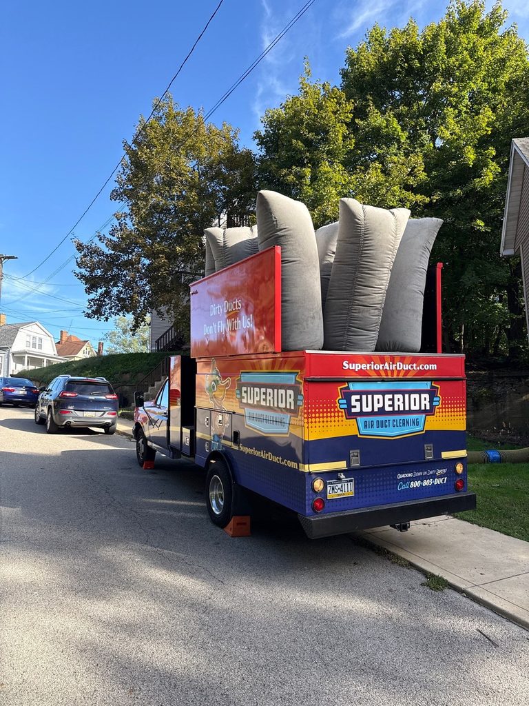 Rear of a Superior Air Duct Cleaning truck with gray filter bags loaded on top, parked curbside on a sunny residential street